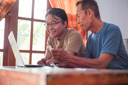 Happy adult son and old father pay online using laptop and credit card. Young and senior togetherness conceptの写真素材