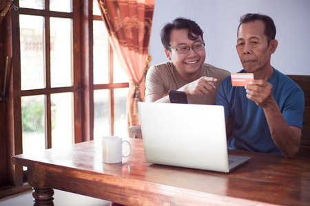 Happy adult son and old father pay online using laptop and credit card. Young and senior togetherness conceptの写真素材