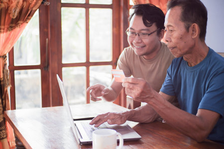 Happy adult son and old father pay online using laptop and credit card. Young and senior togetherness conceptの写真素材
