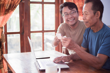 Happy adult son and old father pay online using laptop and credit card. Young and senior togetherness conceptの写真素材