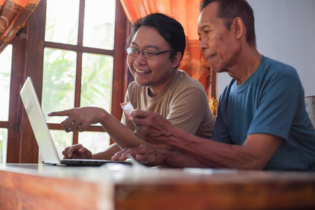 Happy adult son and old father pay online using laptop and credit card. Young and senior togetherness conceptの写真素材