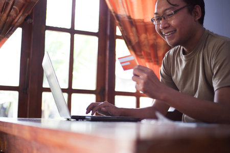 Asian Male with shirt doing online transaction in living room with credit card smartphone and laptop. Freelancer work at home conceptの写真素材