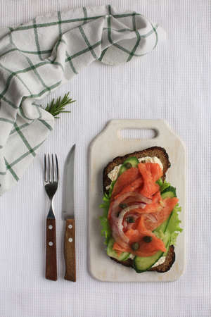 Big tasty Smorrebrod with salmon, green vegatbles, onion, capers, cucumber, white cheese, pepper and rye bread laying on white towel on wooden board. Fork and knife with wooden handles are nearby.の写真素材