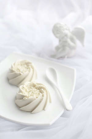 Light airy dessert panna cotta mousse with white chocolate and vanilla on top, on white ceramic plate and ceramic spoon and porcelain angel statuette. Monochromeの写真素材