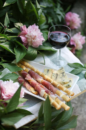 Apperitive with a glass of red wine, fresh prosciutto, grissini sticks and cheese near peony flowers in summer.の写真素材
