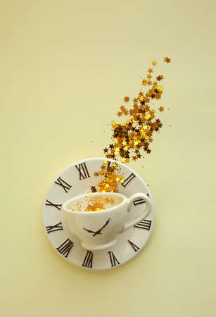 A cup of coffee with tinsel and decorating golden stars inside, placed on small plate with clock...