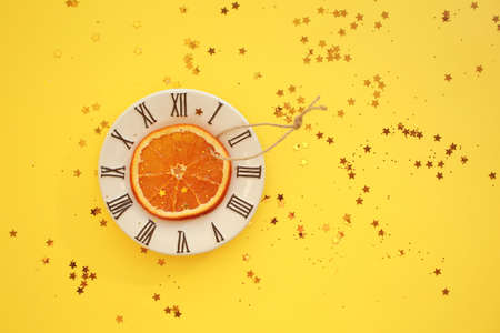 One dryed orange slice on small plate with clock face icon and glitter stars. Bright yellow background.の写真素材