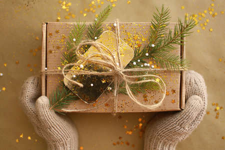 Christmas present holding by two hands in mittens, Craft paper box decorated with evergreen tree, golden card and cord on paper background.の写真素材