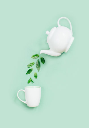 top view of a white teapot pouring tea leaves into a white cup on a light green backgroundの写真素材