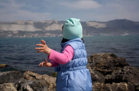 Picture from the back of a little girl running away to the sea and big stones on the shore. A girl is in a blue jacket, cap.の写真素材