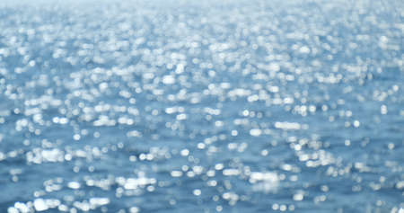 Blurred blue sea sun glare reflections on surface, close-up. Summer defocus background. Sunshine flicker bokeh on seawater. Shine of marine waves motion. Abstract sparkling blurry textureの写真素材
