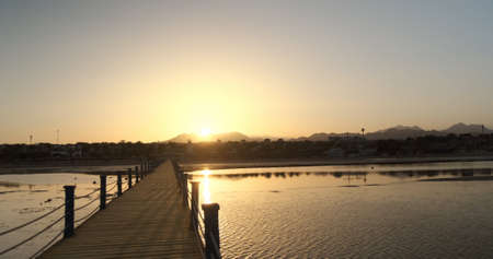 Orange sunset on wooden pier, mountain views, resort hotel, palm trees, beach. Sun sets behind mountain silhouettes, reflected in sea ripples of low tide. Vacation by sea, calm summer landscapeの写真素材
