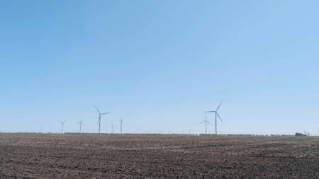 Modern windmills for energy production on plowed field against blue sky on sunny day. Wind turbines generating clean renewable energy. Alternative energy extreme wide shot. No peopleの写真素材