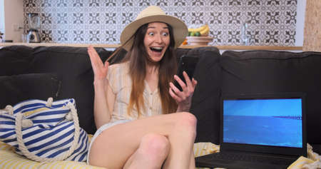 Happy woman rejoices at quarantine ending, opportunity to travel. Excited girl traveler surfing on phone, sitting on couch yellow mat, near beach bag, sea video laptop. Concept finding cheap ticketsの写真素材