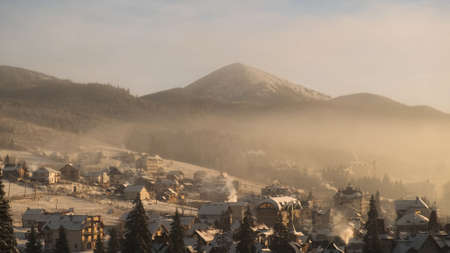 View of ski resort hotels in winter morning. Magnificent view of village in mountains, roofs of houses covered with snow, smoke coming from chimneys. Snow-capped mountains and pine forest in Bukovelの写真素材