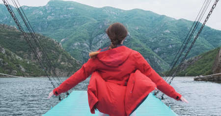 Traveler in red jacket raises arms to sides on moving boat, background of mountains landscape. Woman...