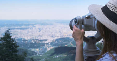 Unrecognizable woman traveler looking through viewing binoculars at summer city. Copy space. Woman in straw hat observes of blur panorama, with binoculars at observation deck. Mountain Dajti Albaniaの写真素材