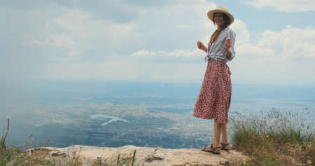 Happy traveler on mountain top. Carefree woman traveler in hat, red dress dancing, jumping on mountain, backdrop of Tirana city, Albania. Happy person enjoying travel by country. Concept of wanderlustの写真素材