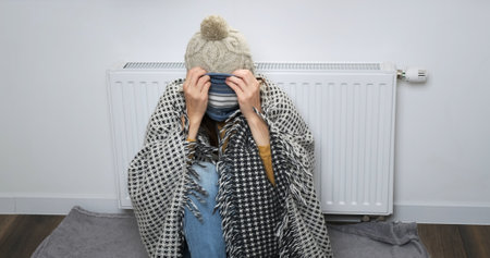 Funny woman in warm clothes freezing at home. Girl puts on hat in white room, pulls scarf over her nose, freezes indoors. Woman backdrop of central heating battery trying to warm herselfの写真素材