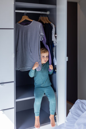 Little smiling boy sitting in the wardrobe and playing in hide and seek. Adorable child is playing at home in the wardrobe.の写真素材