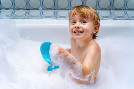 Sweet blond toddler boy playing with toy by taking bath in bathtub. Happy kid smiling and looking at the camera. Carefree childhood.の写真素材