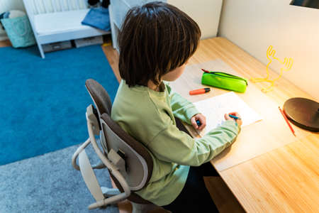 Young school boy doing his homework sitting on orthopedic chair at the table at home. Concentrated kid writing exercises. Homeschooling concept.の写真素材