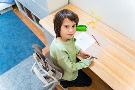 Young school boy doing his homework sitting on orthopedic chair at the table at home. Concentrated kid writing exercises with pleasure. Homeschooling concept.の写真素材
