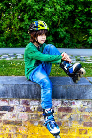 Young boy wearing jeans resting after roller blading, sitting on the fence in the park, outdoors...