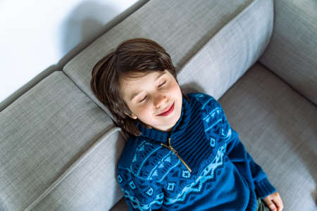 Portrait of young boy sitting on the couch at home, threwing back his head and closing eyes with...