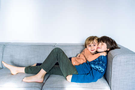 Two brothers lying on the couch at home, smiling and hugging. Good tender relationships between siblings. Embracing children.の写真素材