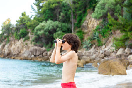 A young boy looking through binoculars staying at the seaside on the beach. Child spending summer...