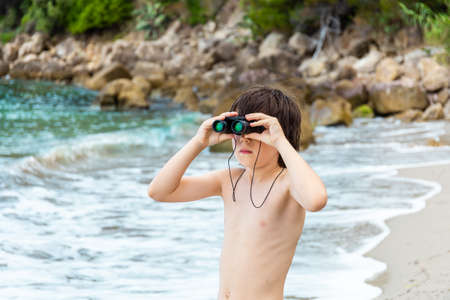 A young boy looking through binoculars staying at the seaside on the beach. Child spending summer holidays at the sea, discovering new horizons.の写真素材