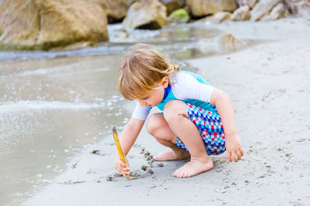Adorable blond toddler boy plays on the beach at the seaside. A young boy drawing with bamboo stick...