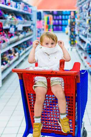 Adorable toddler boy sitting in a shopping cart in a supermarket, putting on protection mask during virus outbreak. Covid epidemic.の写真素材