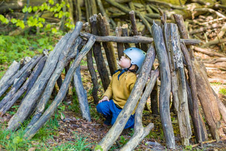 A young boy is playing in the forest in summer or spring. Toddler is building a wooden hut of logs and branches. Child hiding in wigwam created in the park.の写真素材