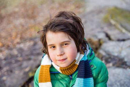 Close uppPortrait of a young boy of nine years old wearing green puffer jacket and a scarf, walking outdoor in a park or forest.の写真素材