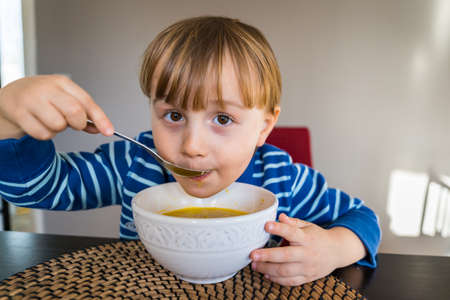 Cute little child having lunch at home. Toddler sitting at the table, tasting soup in a bowl. Happy smiling boy eating healthy domestic food.の写真素材