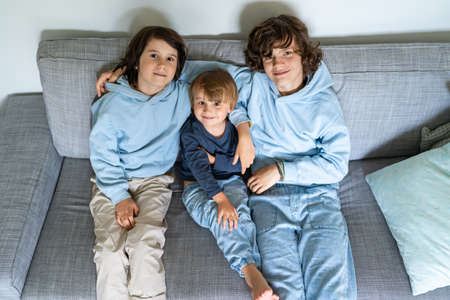 Three brothers of different age are sitting on a sofa at home. The elder boys hug the younger. Siblings good relations.の写真素材