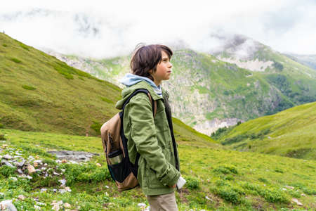 Portrait of a young boy with backpack in a green jacket standing in the mountains in French Alps...