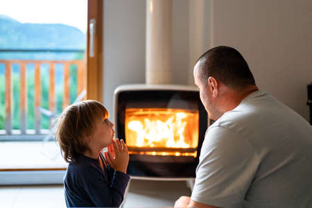 Father and son are sitting by the fireplace in a living-room in a chalet in the mountains. Parents and children concept. Spending time together. Boy and dad at home in the evening.の写真素材