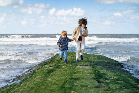 Two brothers holding hands are running on a breakwater at the North Sea. Laughing boys spending weekend at the seaside in Belgium, Knokke. Siblings friendship.の写真素材