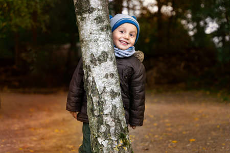 Little boy wearing warm jacket and a hat peeking out from behind a birch trunk. Happy child walking...
