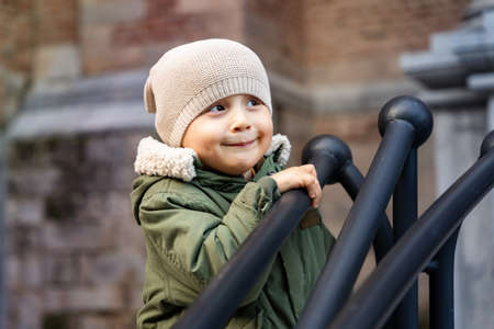 Little boy staying outdoors and holding onto the railing near the building. Child walking in the street. Toddler wearing warm parka and a funny hat looking aside, close up.の写真素材
