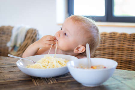 Little boy sitting at the table in a kitchen and eating with pleasure spaghetti with cheese. Toddler taking lunch. Healthy simple food for children concept.の写真素材