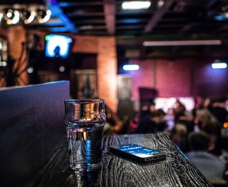 Close-up of a smart phone and a glass of water on a desk in bar. Evening in bar