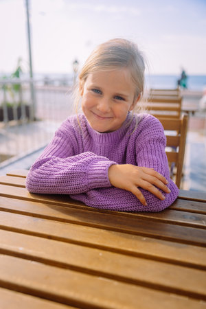 In a good mood. Happy little girl sitting at cafe outdoors holding her hands as at the desc, cute smiling girl portrait with copy space. High quality photoの写真素材