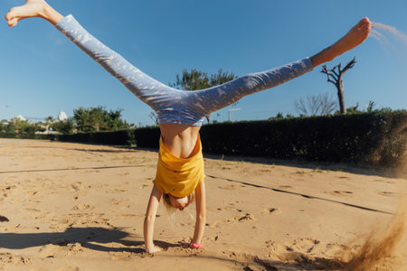 Little girl stands on hands in sand on a sunny day, girl wearing yellow t-shirt and bleu leggings...