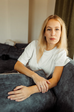 Serious blonde Caucasian girl sitting on sofa and looking curiously at camera. High quality photoの写真素材