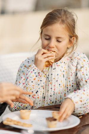 Cute little girl eating tasty crepes in cafe outdoors feeling satisfied and hungry with closed eyes. High quality photoの写真素材