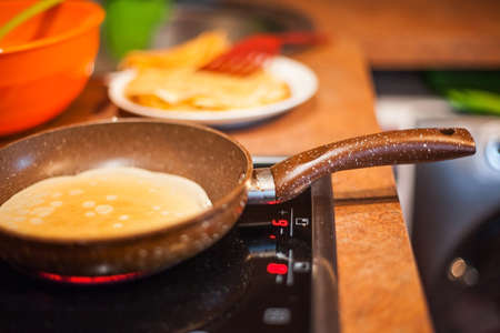 Plain thin pancake frying on a brown pan placed on electric cooker in a warm light, colorful utensils on background, low depth of fieldの写真素材